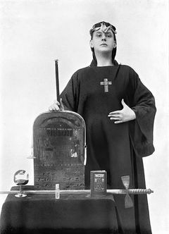 Aleister Crowley em seu Robe e Coroa, armado com Baqueta, Taça, Espada, Pantáculo, Sino, Livro e Óleo Santo.